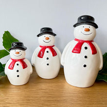 A set of three ceramic snowmen of varying sizes with hand-painted details, wearing red scarves and black hats, positioned on a wooden surface.