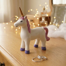 A small white ceramic unicorn figurine with a pink mane and tail, purple hooves, and a gold horn, standing on a lavender surface with two silver rings in front of it.
