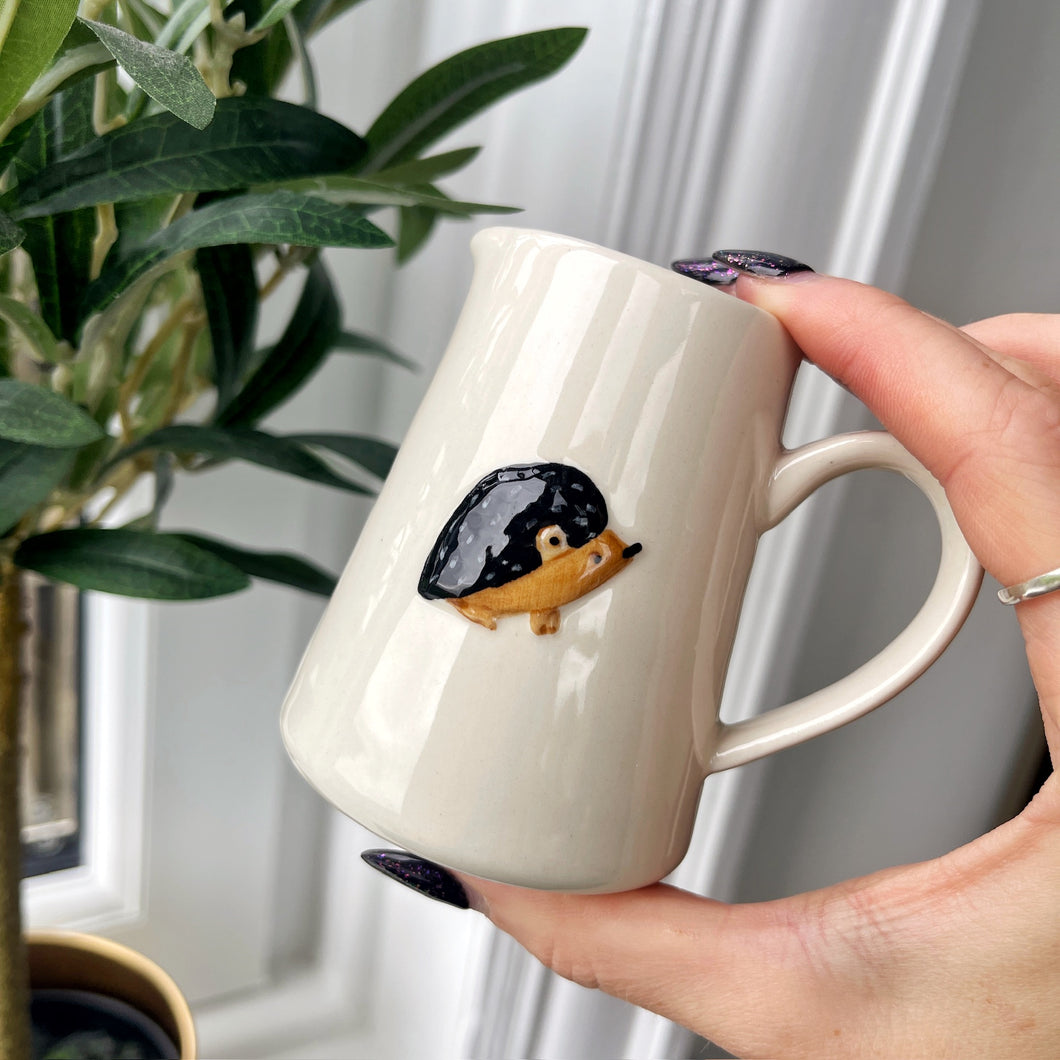 Person holding a white mug with a hedgehog design in front of a plant