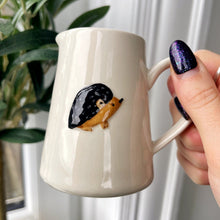 Person holding a white mug with a hedgehog design in front of a plant.
