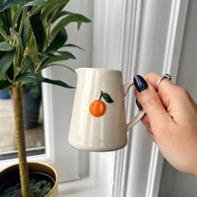Hand holding a white mug with an orange design next to a potted plant.