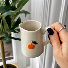Hand holding a white mug with an orange design in front of a window with curtains and a plant.