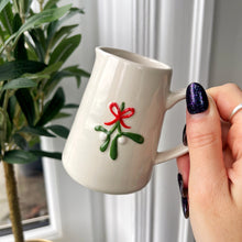 White mug with embroidered mistletoe design held by a hand, with a plant in the background.