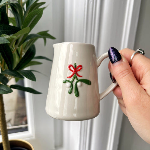 White mug with a red bow design held by a hand in front of a window with white curtains.