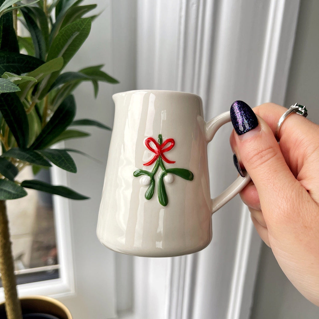 White mug with a red bow design held by a hand in front of a window with white curtains.