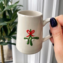 White mug with a decorative mistletoe design held by a hand, with a plant in the background.