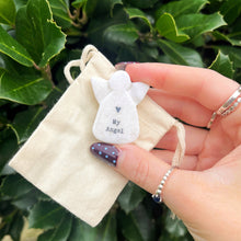Hand holding a small angel figurine with 'My Angel' text on a beige pouch against a green leafy background.