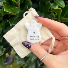 Hand holding a small white angel figurine with 'Watching over you' text on a beige fabric pouch against a green leafy background.