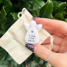 Hand holding a small white angel figurine with 'A hug from heaven' text on a beige fabric pouch against a green leafy background.
