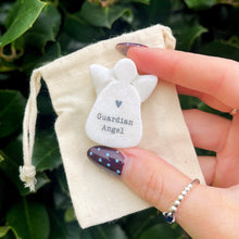Hand holding a small white bag with a 'Guardian Angel' charm against a green leafy background