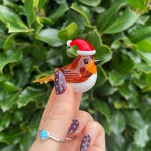 Glass robin figurine wearing a Santa hat held against a green leafy background