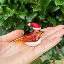 Glass robin figurine wearing a Santa hat held in a hand with a green leafy background