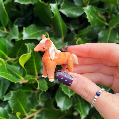 Hand holding a small orange Horse figurine against a green leafy background