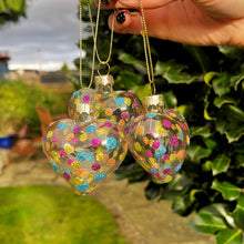 Hand holding two clear heart-shaped decorations with colorful confetti against a garden background