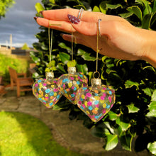 Hand holding three colorful heart-shaped ornaments against a garden background