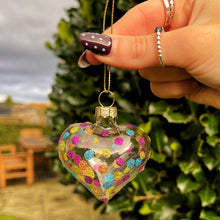 Heart-shaped glass ornament with colorful decorations held by a hand outdoors.
