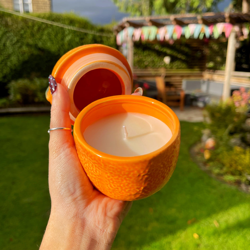 Hand holding an orange container with a candle inside, set against a garden background.