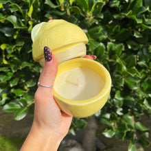 Yellow lemon-shaped candle held by a hand against a green leafy background