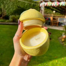 Yellow lemon-shaped candle held by a hand outdoors with greenery in the background