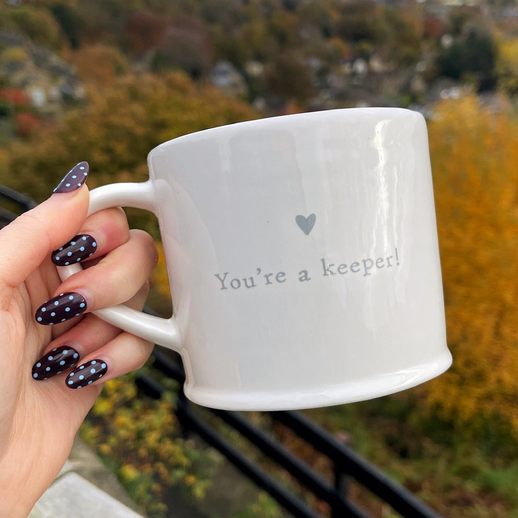 Hand holding a mug with 'You're a keeper' text against an autumn landscape.