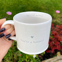 White mug with 'Special friend You're a keeper!' text held by a hand against a garden background