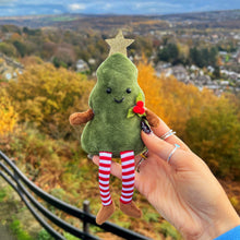 Hand holding a green plush toy with a star on top, striped socks, and a red flower against an autumn landscape.