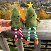 Two green plush Christmas trees with star hats and striped legs sitting on a wooden railing with a scenic background.