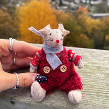 Small plush toy mouse in a red outfit with buttons, held by a hand against a scenic background.