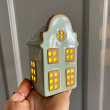 Ceramic house-shaped lantern with glowing windows held by a hand against a neutral background