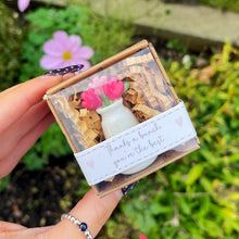 Hand holding a small wooden box with floral design and text, against a natural background with flowers and greenery.