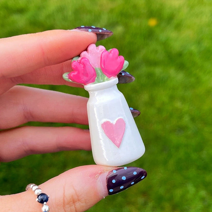 Hand holding a small white bottle with pink flowers and a heart on a grassy background
