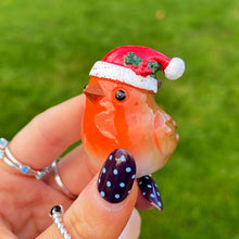 Hand holding a small figurine of a Robin wearing a Santa hat against a grassy background