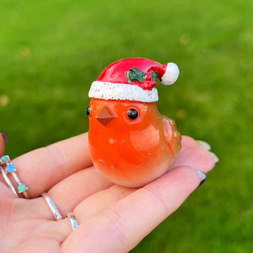 Small orange bird figurine wearing a Santa hat held in a hand with a grassy background.