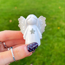 White angel figurine held by a hand with painted nails against a grassy background.