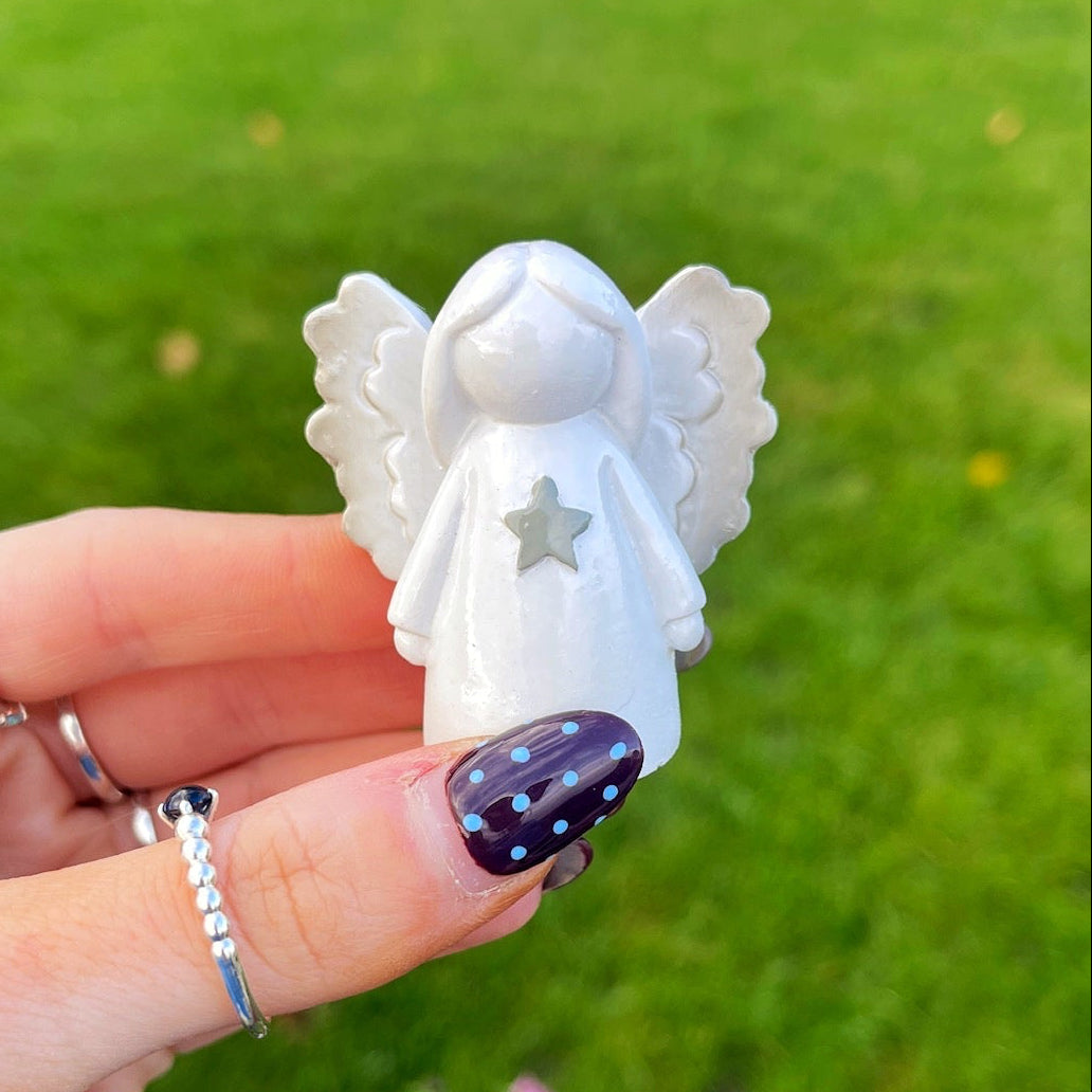 White angel figurine held by a hand with painted nails against a grassy background.