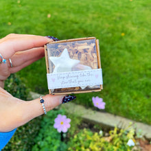 Hand holding a small wooden box with a star-shaped object and a message card against a green outdoor background.