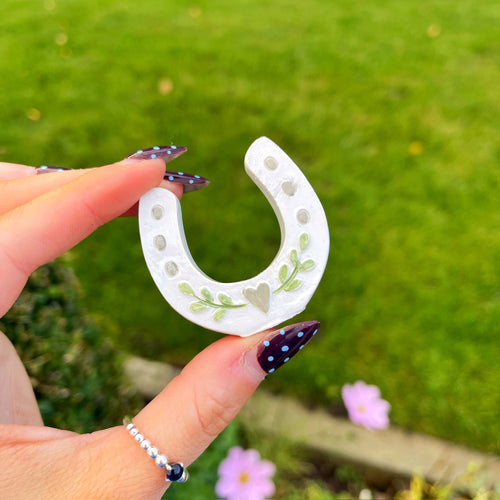 Hand holding a decorative white horseshoe with green patterns against a green outdoor background.