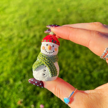 Hand holding a small snowman figurine with a red hat and green scarf against a grassy background