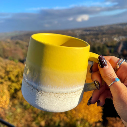 Hand holding a yellow and white mug with a scenic background
