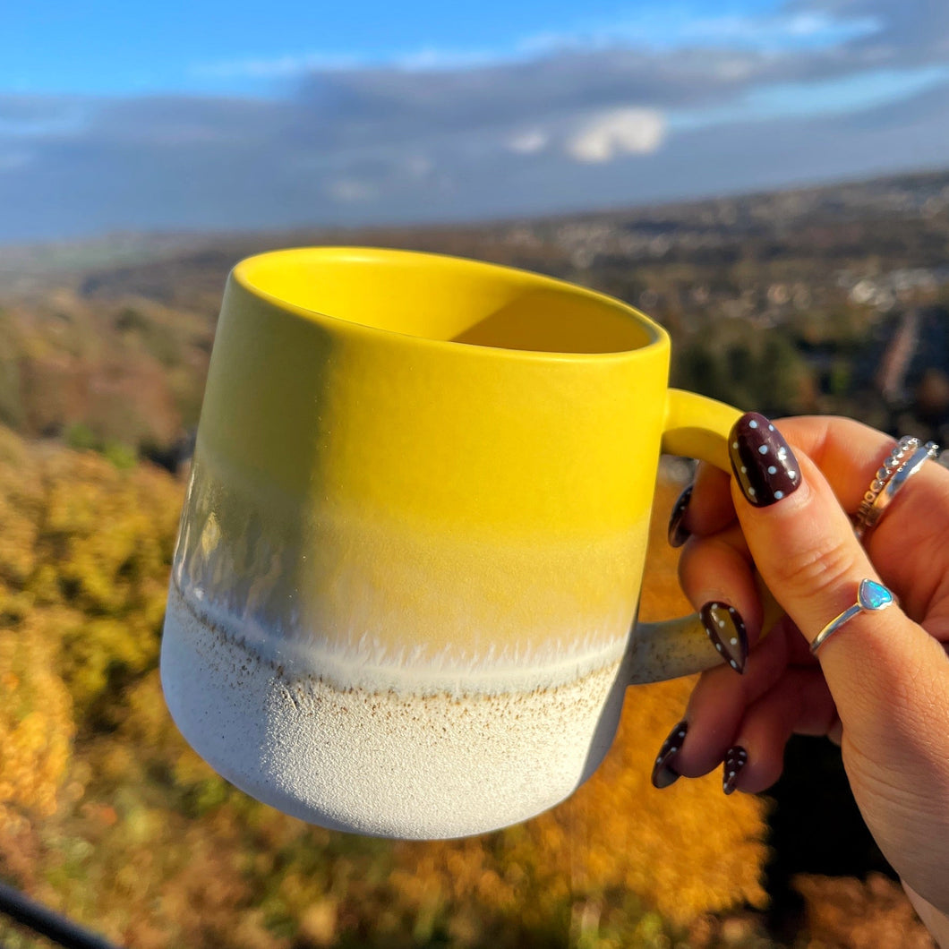 Hand holding a yellow and white mug with a scenic background