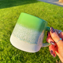 Hand holding a green mug with a gradient effect against a grassy outdoor background