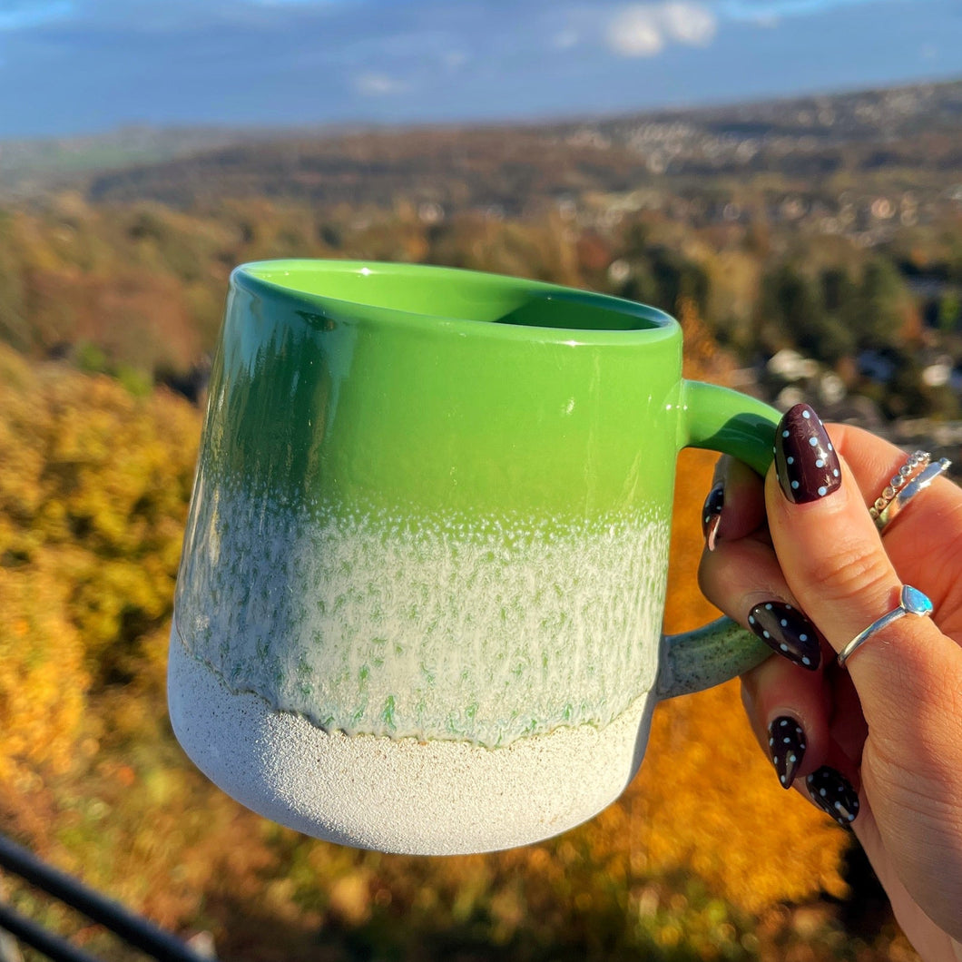 Green and white mug held by a hand with a scenic background of trees and hills.