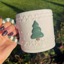 White mug with a textured pattern and green tree design held by a hand outdoors.