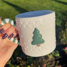 White mug with blue interior, textured surface, and green tree design held outdoors.