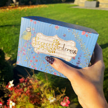 Hand holding a 'Secret Garden' box with floral design outdoors