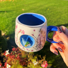Person holding a decorative mug with a blue interior and bird design outdoors.