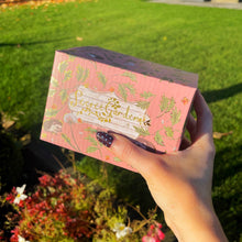 Hand holding a pink box with floral design against a garden background