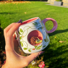 Hand holding a decorative mug with floral patterns and a small hedgehog design outdoors.