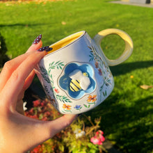 Hand holding a decorative mug with a bee design against a garden background