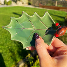 Hand holding a green leaf-shaped object with red berries against a blurred outdoor background